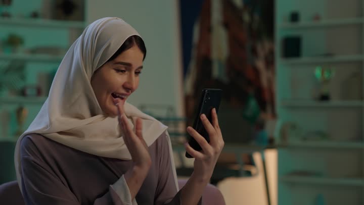 اجراء مكالمة فيديو، رفع كف اليد بتعابير التحية و السلام، الدردشة والتواصل مع الاهل والاصدقاء، استخدام الاجهزة التقنية الحديثة، دمج التكنولوجيا المتطورة في الحياة اليومية، شابة عربية خليجية سعودية محجبة ترتدي العباءة  تحمل بيدها الهاتف المحمول