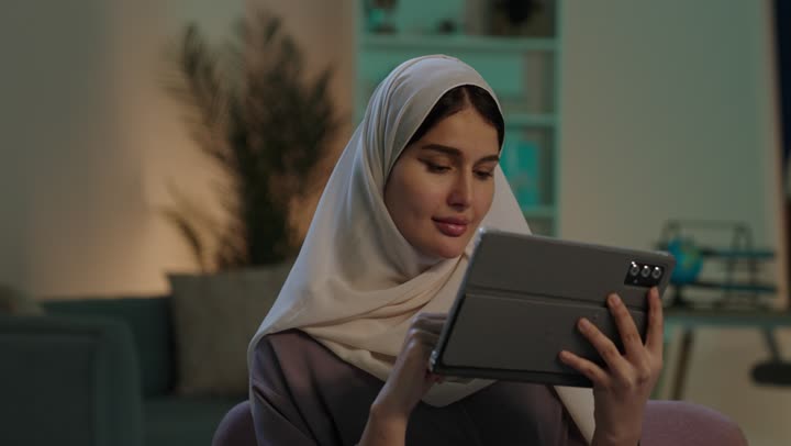 النظر للكاميرا بتعابير السعادة و السرور، استخدام الاجهزة التقنية الحديثة، الدردشة والتواصل مع الاهل والاصدقاء، دمج التكنولوجيا المتطورة في الحياة اليومية، شابة عربية خليجية سعودية محجبة ترتدي العباءة  تحمل بيدها جهاز التابلت