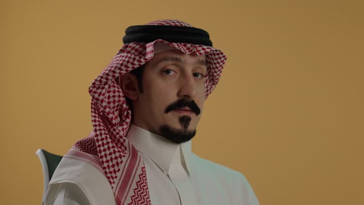 حركة أصبع تدل على القبول والرضا، الإمتياز والتميز والنجاح، بورتريه مقرب لرجل عربي خليجي سعودي يرتدي الثوب الابيض و الشماغ يجلس على كرسي يرفع أصبع الإبهام لأعلى، إيماءات السعادة والإعجاب والموافقة، خلفية بنية