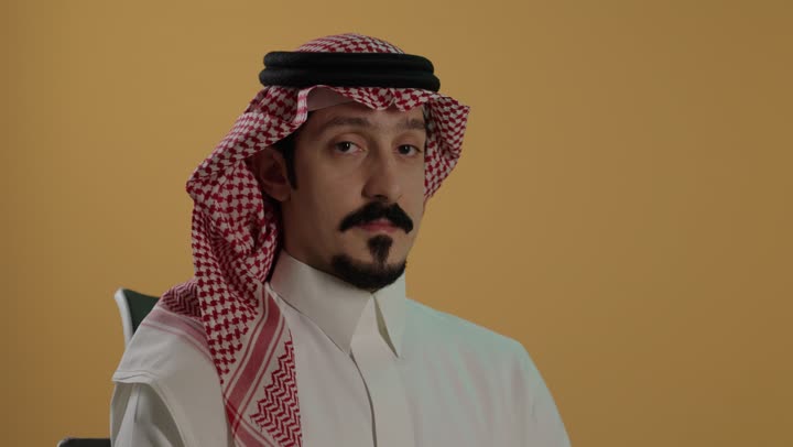 حركة يد وايماءات عدم الإعجاب والموافقة،بورتريه مقرب لرجل عربي خليجي سعودي يرتدي الثوب الابيض و الشماغ يجلس على كرسي يشير بايماءات عدم القبول، الإشارة بإصبع الإبهام للاسفل دليل الرفض، خلفية بنية