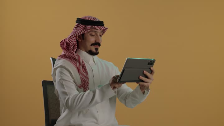 The use of advanced technology, browsing social media, communicating and chatting with relatives, a portrait of a Saudi Arabian Gulf man wearing a white thobe and a ghutrah sitting on a chair holding a tablet device, brown background.