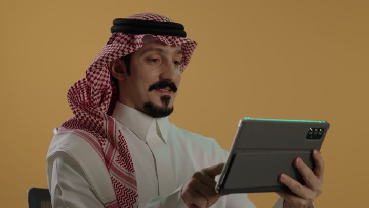 تصفح مواقع التواصل الاجتماعي، التواصل و الدردشة مع الاقارب، استخدام التكنولوجيا المتطورة , بورتريه لرجل عربي خليجي سعودي يرتدي الثوب الابيض و الشماغ يجلس على كرسي يحمل بيده جهاز التابلت اللوحي، خلفية بنية