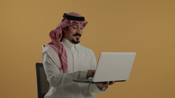 The concept of remote work, typing on a keyboard, a portrait of an Arab Gulf Saudi man wearing a white thobe and a ghutrah sitting on a chair using a laptop, brown background.