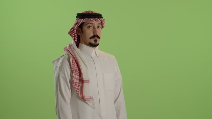 Standing with crossed arms, elegance and attention to appearance, a portrait of a Saudi Arabian Gulf man wearing a white thobe and a ghutrah, looking at the camera with gestures of self-confidence, green background.