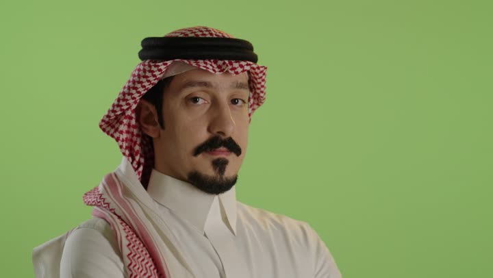 الاناقة و الاهتمام بالمظهر الخارجي ، الوقوف بايدي متقاطعة ، بورتريه مقرب لرجل عربي خليجي سعودي يرتدي الثوب الابيض و الشماغ ينظر الى الكاميرا بايماءات الثقة بالنفس ، خلفية خضراء