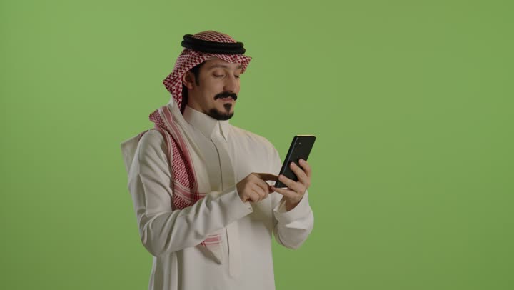 استخدام التقنيات الحديثة، الدردشة عبر الانترنت ، تصفح مواقع التواصل الاجتماعي عبر الجوال، إنجاز مهام العمل عن بعد، بورترية لرجل عربي خليجي سعودي يرتدي الثوب الابيض و الشماغ يقوم باستخدام الهاتف المحمول، خلفية خضراء
