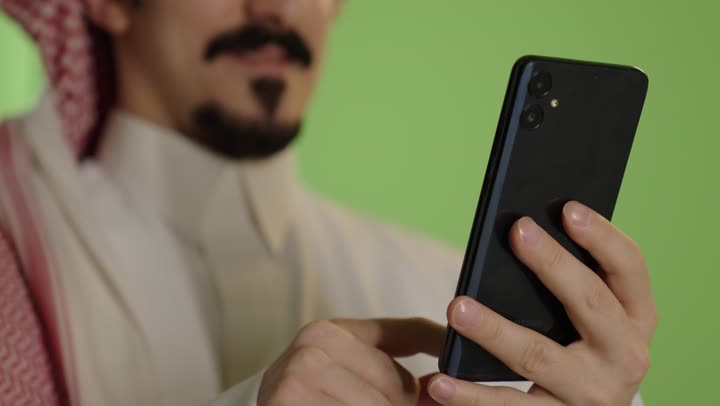 Online chatting, using modern technologies, browsing social media via mobile, completing work tasks remotely, a close-up portrait of the hand of an Arab Gulf Saudi man wearing a white thobe and a ghutrah using a mobile phone, green background.