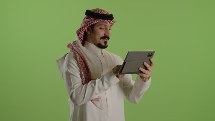 النظر للكاميرا بتعابير السرور، الدردشة والتواصل مع الأهل والأصدقاء، تصفح مواقع التواصل الاجتماعي، استخدام التكنولوجيا المتطورة , بورتريه لرجل عربي خليجي سعودي يرتدي الثوب الابيض و الشماغ يحمل بيده جهاز التابلت اللوحي، خضراء بنية