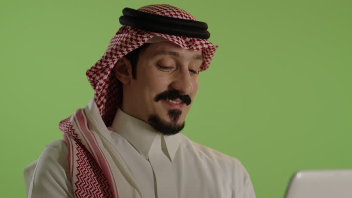 إدارة شؤون الأعمال وتنظيمها عبر الاجهزة التقنية، مهنة ووظيفة ادارية، دمج العمل مع التكنولوجيا، بورتريه لرجل عربي خليجي سعودي يرتدي الثوب التقليدي والشماغ يعمل على الحاسوب المحمول، استخدام جهاز تقني حديث ومتطور، العمل عن بعد وانجاز المهام المطلوبة، خلفية خضراء