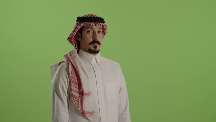 النظر إلى الكاميرا بإيماءات السعادة والسرور، مفهوم الاعجاب والرضا، بورتريه لرجل عربي خليجي سعودي يرتدي الثوب الابيض والشماغ يجلس على كرسي يرفع اصبع الإبهام نحو الأعلى بتعابير القبول والموافقة، خلفية خضراء