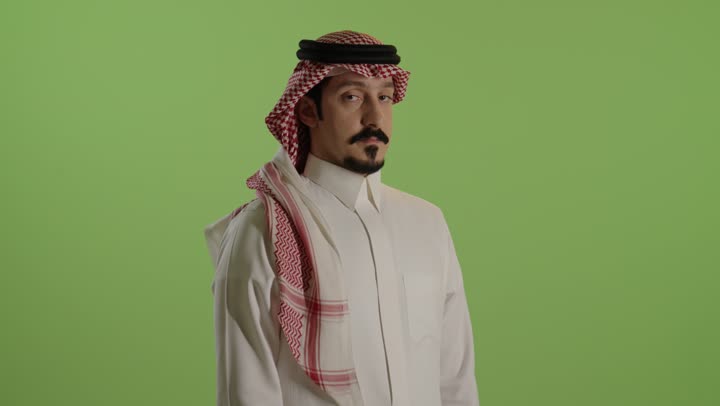 The concept of negative evaluation and discomfort, anxiety, and failure to achieve something, a portrait of a Saudi Arabian Gulf Arab man wearing a white thobe and a ghutrah looking at the camera, pointing his thumb down with gestures of discomfort, expressions of rejection, non-acceptance, and dissatisfaction, green background.