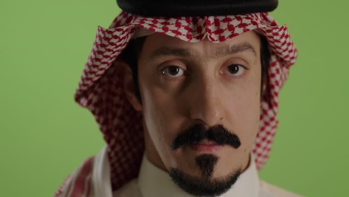 التوقف عن الكلام وخفض الصوت، الهدوء والسكون وعدم الإزعاج، بورتريه مقرب لرجل عربي خليجي سعودي يرتدي الثوب الابيض والشماغ يضع أصبع السبابة على فمه، طلب السكوت والصمت، حركة أصبع وإشارات، خلفية خضراء