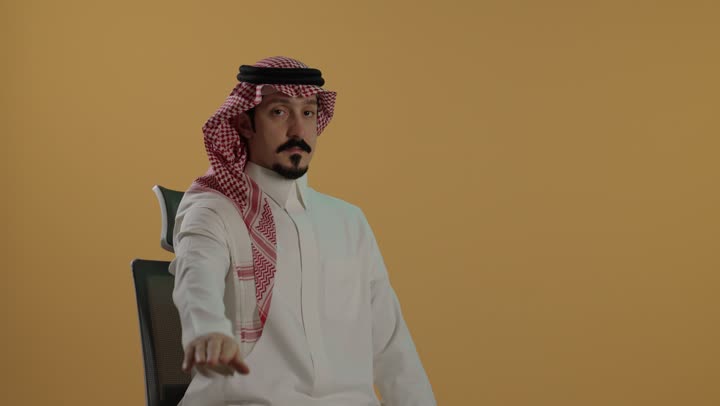 مفهوم الرفض والامتناع والانزعاج, بورتريه لرجل عربي خليجي سعودي يرتدي الثوب الابيض والشماغ يمد يده نحو الأمام مع رفع الكف بإيماءات النهي عن فعل امر ما, طلب التوقف وعدم الاقتراب, تعابير الحزم والجدية, خلفية بنية