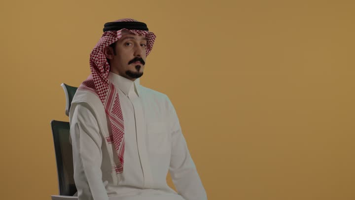 مفهوم الاعجاب والرضا، النظر إلى الكاميرا بإيماءات السعادة والسرور، بورتريه لرجل عربي خليجي سعودي يرتدي الثوب الابيض والشماغ  يجلس على كرسي يرفع اصبع الإبهام نحو الأعلى بتعابير القبول والموافقة، خلفية بنية