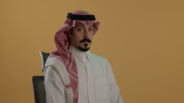 طلب السكوت والصمت، الهدوء والسكون وعدم الإزعاج، بورتريه مقرب لرجل عربي خليجي سعودي يرتدي الثوب الابيض والشماغ يضع أصبع السبابة على فمه، التوقف عن الكلام وخفض الصوت، خلفية بنية