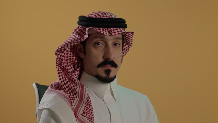 الهدوء والسكون وعدم الإزعاج، بورتريه مقرب لرجل عربي خليجي سعودي يرتدي الثوب الابيض والشماغ يضع أصبع السبابة على فمه، التوقف عن الكلام وخفض الصوت، طلب السكوت والصمت، حركة أصبع وإشارات، خلفية بنية