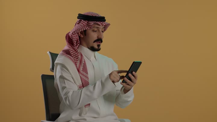 Using a modern and advanced technical device, remote work, browsing applications and programs, social media communication online, a portrait of an Arab Gulf Saudi man wearing a white thobe and a ghutrah sitting on a chair holding a mobile phone, brown background.
