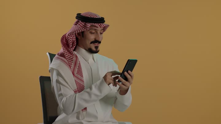 Remote work, browsing applications and programs, social media communication online, using a modern and advanced technical device, a portrait of a Saudi Arabian Gulf man wearing a white thobe and a ghutrah sitting on a chair holding a mobile phone, brown background.
