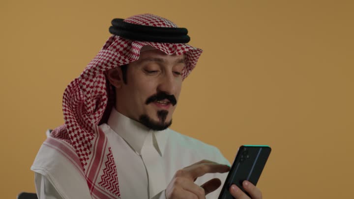 Browsing applications and programs, online social communication, remote work, using a modern and advanced technical device, a portrait of a Saudi Arabian Gulf man wearing a white thobe and a ghutrah sitting on a chair holding a mobile phone, brown background.