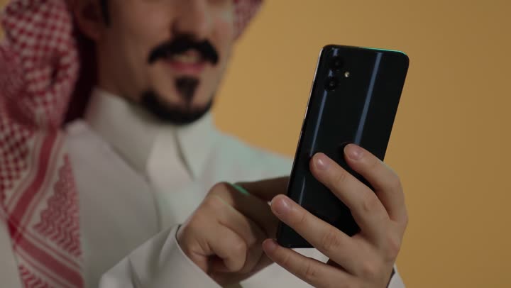 Online social communication, browsing applications and programs, remote work, using a modern and advanced technical device, a close-up portrait of a Saudi Arabian Gulf man wearing a white thobe and a ghutrah sitting on a chair holding a mobile phone, brown background.