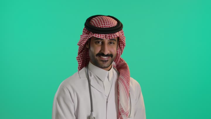 Medical examination and providing treatment services to patients, a portrait of an Arab Gulf Saudi doctor wearing traditional attire and a ghutrah, holding a stethoscope, standing with arms crossed, looking up with gestures of self-confidence, green background, chroma.
