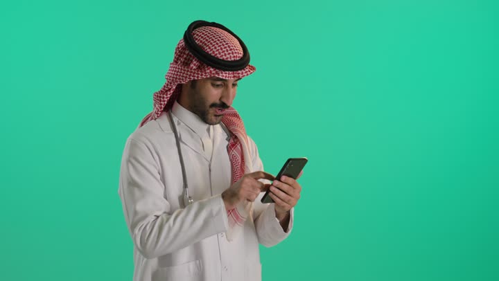 استخدام الأجهزة التقنية الحديثة ودمج التكنولوجيا مع الطب، التطبيقات الرقمية للخدمات الصحية, تقديم الخدمات والرعاية الصحية عبر الإنترنت، بورتريه لطبيب عربي خليجي سعودي يرتدي الثوب و الشماغ و المعطف الطبي و السماعه يقوم باستخدام الهاتف المحمول، خلفية خضراء ، كروما
