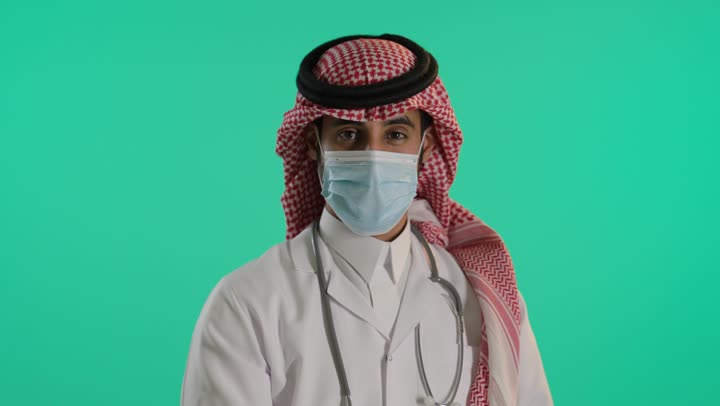 Following COVID-19 prevention measures, safety and protection from the coronavirus, a portrait of an Arab Gulf Saudi doctor wearing traditional attire, gloves, and a mask to control the spread of infection, with a stethoscope standing with arms crossed, green background, chroma.