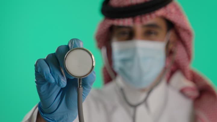 مفهوم الطب والرعاية الصحية،ارتداء ماسك كورونا للوقاية، إجراء الفحوصات اللازمة، بورتريه مقرب لطبيب عربي خليجي سعودي يرتدي الزي التقليدي و الشماغ  والمعطف الطبي، استخدام سماعة الطبيب لسماع نبضات القلب، العمل في القطاع الصحي السعودي، خلفية خضراء، كروما