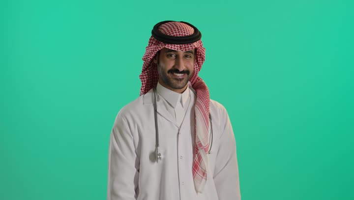 Medical examination and providing treatment services to patients, a portrait of an Arab Gulf Saudi doctor wearing traditional attire and a ghutrah, holding a stethoscope, standing with arms crossed, looking up with gestures of self-confidence, green background, chroma.