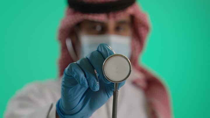 إجراء الفحوصات اللازمة، مفهوم الطب والرعاية الصحية،ارتداء ماسك كورونا للوقاية، بورتريه مقرب لطبيب عربي خليجي سعودي يرتدي الزي التقليدي و الشماغ  والمعطف الطبي، استخدام سماعة الطبيب لسماع نبضات القلب، العمل في القطاع الصحي السعودي، خلفية خضراء، كروما