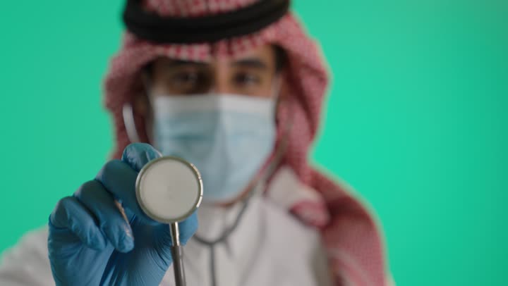 Wearing a coronavirus mask for protection, conducting necessary tests, the concept of medicine and healthcare, a close-up portrait of an Arab Gulf Saudi doctor wearing traditional attire and a shemagh and a medical coat, using a stethoscope to listen to heartbeats, working in the Saudi health sector, green background, chroma.