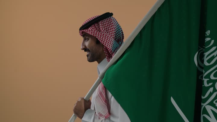 شعار الهوية الوطنية وراية الإسلام والتوحيد، ذكرى تأسيس الدولة السعودية الأولى 22 فبراير، النظر الى الكاميرا بتعابير السرور، الفخر والاعتزاز بإنجازات الوطن، بورتريه لرجل عربي خليجي سعودي يرتدي الثوب و الشماغ يحمل علم التأسيس، خلفية بنية