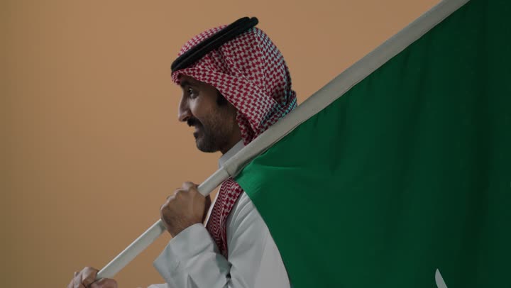 ذكرى تأسيس الدولة السعودية الأولى 22 فبراير، النظر الى الكاميرا بتعابير السرور، شعار الهوية الوطنية وراية الإسلام والتوحيد، الفخر والاعتزاز بإنجازات الوطن، بورتريه لرجل عربي خليجي سعودي يرتدي الثوب و الشماغ يحمل علم التأسيس، خلفية بنية