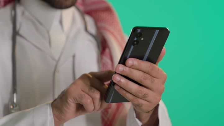 تقديم الخدمات العلاجية، استخدام التقنية الحديثه في المجال الطبي, بورتريه مقرب ليد طبيب عربي خليجي سعودي يرتدي الثوب الابيض و الشماغ والمعطف الطبي يضع السماعة الطبية يستخدم الهاتف المحمول ، خلفية خضراء ، كروما