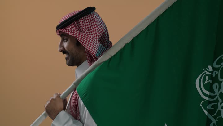 النظر الى الكاميرا بتعابير السرور، ذكرى تأسيس الدولة السعودية الأولى 22 فبراير، شعار الهوية الوطنية وراية الإسلام والتوحيد، الفخر والاعتزاز بإنجازات الوطن، بورتريه لرجل عربي خليجي سعودي يرتدي الثوب و الشماغ يحمل علم التأسيس، خلفية بنية