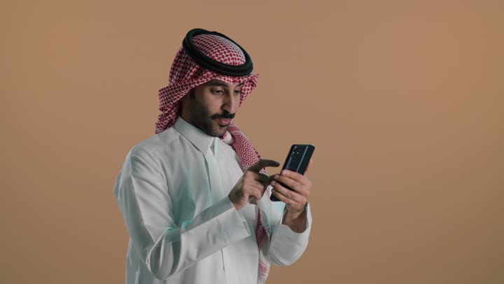 إنجاز مهام العمل عن بعد، استخدام التقنيات الحديثة، الدردشة عبر الانترنت ، تصفح مواقع التواصل الاجتماعي عبر الجوال، بورتريه لشاب عربي خليجي سعودي يرتدي الثوب الابيض و الشماغ يقوم باستخدام الهاتف المحمول، خلفية خضراء، كروما