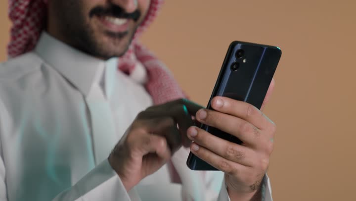 The use of modern technologies, chatting online, browsing social media on mobile, completing work tasks remotely, a close-up portrait of a young Arab Gulf Saudi man wearing a white thobe and a ghutrah using a mobile phone, green background, chroma.