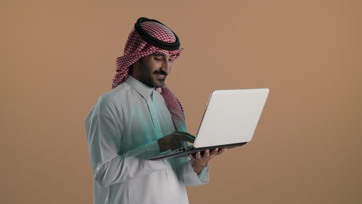 إدارة شؤون الأعمال وتنظيمها عبر الاجهزة التقنية، مهنة ووظيفة ادارية، دمج العمل مع التكنولوجيا، بورتريه لشاب عربي خليجي سعودي يرتدي الثوب التقليدي والشماغ يعمل على الحاسوب المحمول، استخدام جهاز تقني حديث ومتطور، العمل عن بعد وانجاز المهام المطلوبة، خلفية بنية