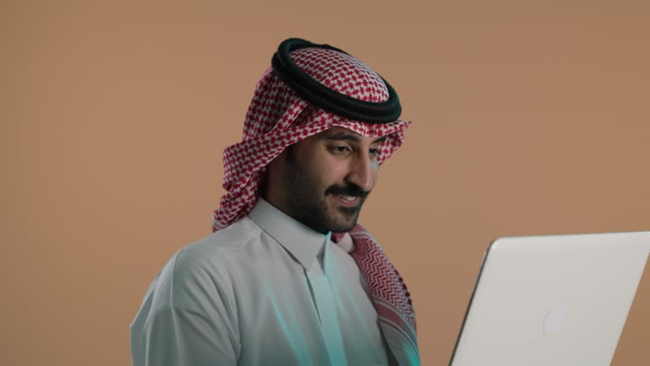 النظر للكاميرا بتعابير السرور، إدارة شؤون الأعمال وتنظيمها عبر الاجهزة التقنية، دمج العمل مع التكنولوجيا، بورتريه لشاب عربي خليجي سعودي يرتدي الثوب التقليدي والشماغ يعمل على الحاسوب المحمول، استخدام جهاز تقني حديث ومتطور، العمل عن بعد وانجاز المهام المطلوبة، خلفية بنية
