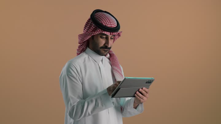 التواصل و الدردشة مع الاقارب، تصفح مواقع التواصل الاجتماعي، استخدام التكنولوجيا المتطورة , بورتريه لشاب عربي خليجي سعودي يرتدي الثوب الابيض و الشماغ  يحمل بيده جهاز التابلت اللوحي، خلفية بنية