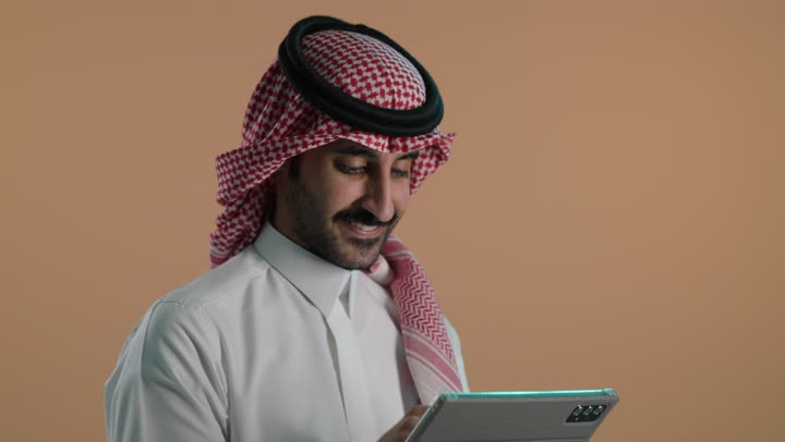 Chatting and communicating with family and friends, browsing social media, using advanced technology, a portrait of a young Arab Gulf Saudi man wearing a white thobe and a ghutrah, holding a tablet device in his hand, with a brown background.