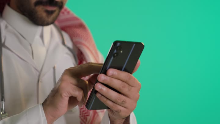 الدردشة عبر الانترنت ، استخدام التقنيات الحديثة، تصفح مواقع التواصل الاجتماعي عبر الجوال، إنجاز مهام العمل عن بعد، بورتريه مقرب ليد شاب عربي خليجي سعودي يرتدي الثوب الابيض و الشماغ يقوم باستخدام الهاتف المحمول، خلفية خضراء، كروما