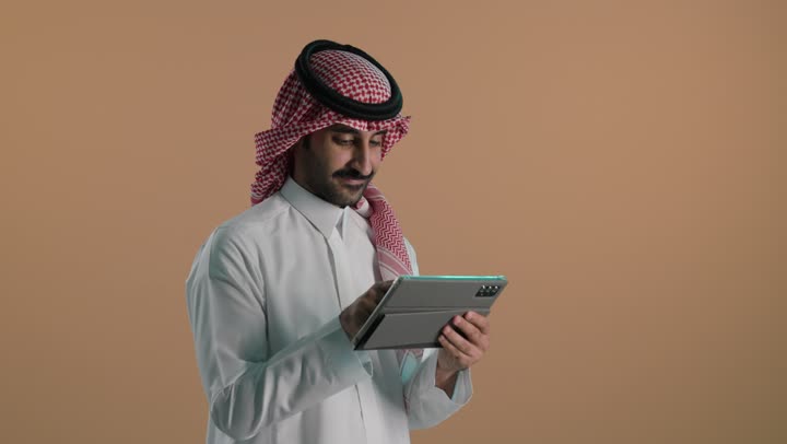 تصفح مواقع التواصل الاجتماعي، استخدام التكنولوجيا المتطورة , بورتريه لشاب عربي خليجي سعودي يرتدي الثوب الابيض و الشماغ  يحمل بيده جهاز التابلت اللوحي، الدردشة والتواصل مع الأهل والأصدقاء، خلفية بنية
