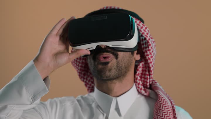 التطور التقني والتكنولوجي، المتعة و التسلية بالمشاهدة عبر نظارة الواقع الافتراضي، تعابير الاندهاش والتفاجؤ, بورتريه لشاب عربي خليجي سعودي  يرتدي الثوب الابيض و الشماغ ينظر من خلال نظارات ثلاثية الأبعاد (الفي ار), الاندماج ومحاكاة العالم الخيالي، خلفية بنية