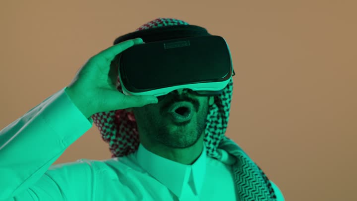 المتعة و التسلية بالمشاهدة عبر نظارة الواقع الافتراضي، تعابير الاندهاش والتفاجؤ، التطور التقني والتكنولوجي, بورتريه لشاب عربي خليجي سعودي  يرتدي الثوب الابيض و الشماغ ينظر من خلال نظارات ثلاثية الأبعاد (الفي ار), الاندماج ومحاكاة العالم الخيالي، خلفية بنية