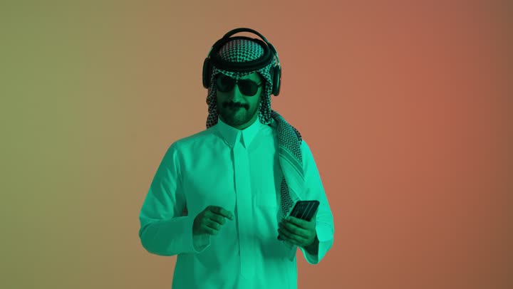 الشعور بالفرح والسعادة، حركات يدين تدل على الرقص والتفاعل مع الموسيقى، بورتريه لشاب عربي خليجي سعودي يرتدي الثوب الابيض و الشماغ و النظارة الشمسية يمسك بيدية الهاتف المحمول يستمتع بسماع الأغاني المتنوعة، خلفية ملونه