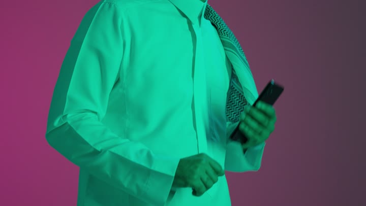 حركات يدين تدل على الرقص والتفاعل مع الموسيقى، بورتريه مقرب لشاب عربي خليجي سعودي يرتدي الثوب الابيض يمسك بيدية الهاتف المحمول يستمتع بسماع الأغاني المتنوعة، الشعور بالفرح والسعادة، خلفية ملونه