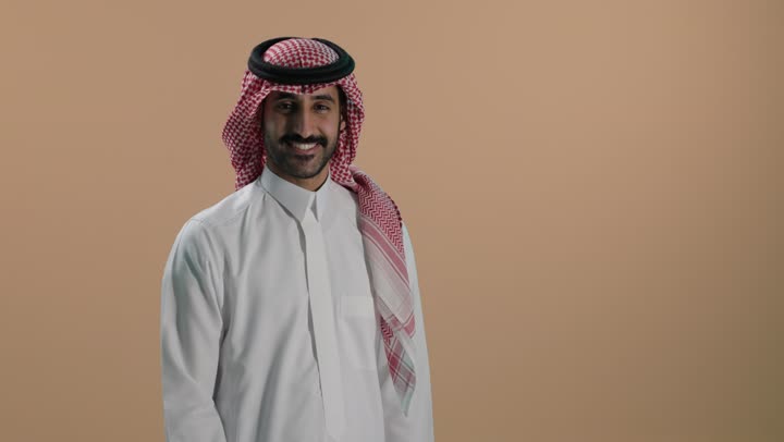 النظر إلى الكاميرا بإيماءات السعادة والسرور، مفهوم الاعجاب والرضا، بورتريه  لشاب عربي خليجي سعودي يرتدي الثوب الابيض والشماغ يرفع اصبع الإبهام نحو الأعلى بتعابير القبول والموافقة، خلفية بنية