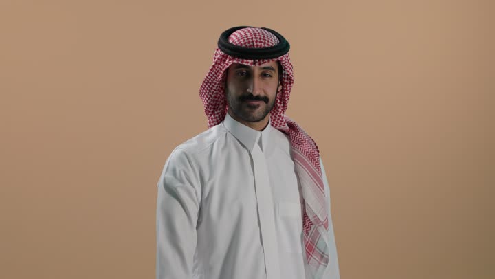 مفهوم الاعجاب والرضا، النظر إلى الكاميرا بإيماءات السعادة والسرور، بورتريه لشاب عربي خليجي سعودي يرتدي الثوب الابيض والشماغ يرفع اصبع الإبهام نحو الأعلى بتعابير القبول والموافقة، خلفية بنية