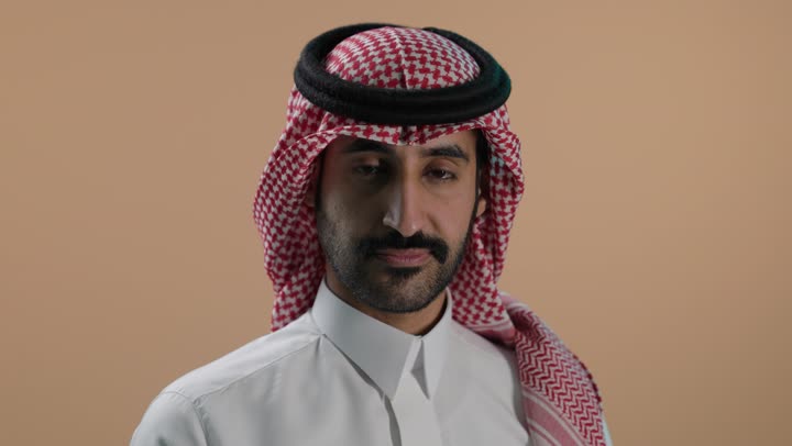 مفهوم التقييم السلبي والانزعاج، القلق والفشل في تحقيق أمر ما، بورتريه مقرب لشاب عربي خليجي سعودي يرتدي الثوب الابيض والشماغ ينظر الى الكاميرا يشير بإصبع الابهام الى الاسفل بإيماءات الانزعاج، تعابير الرفض وعدم القبول والرضا، خلفية بنية